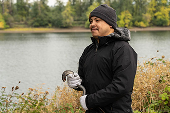 A man in a black jacket and hat stands in front of a river holding a lamprey in gloved hands