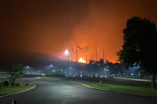 A road under a red sky from a local wildfire. Flames rise above a hill in the background of the road.