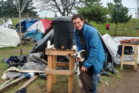 A man holds his dog standing in front of a makeshift handwashing station at a homeless encampment