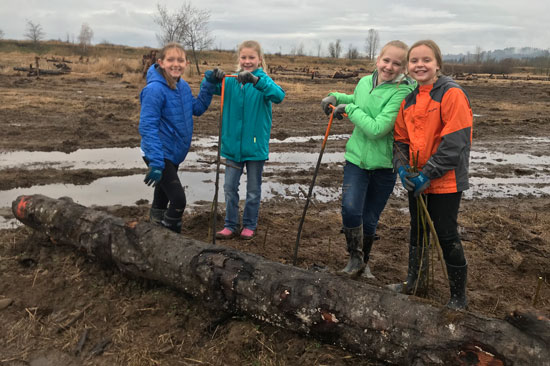 Four kids stand holding digging tools at a muddy restoration site