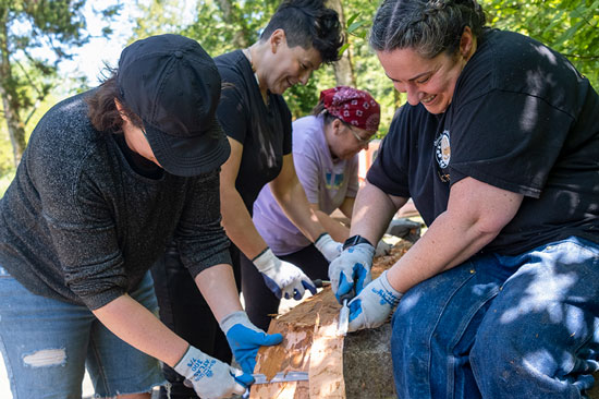 A group of four on either side of a piece of wood strips it using knives