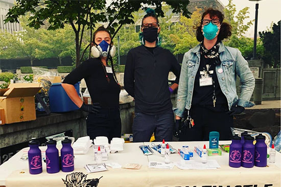 Three people in facemasks stand behind a table that has waterbottles and other supplies on it