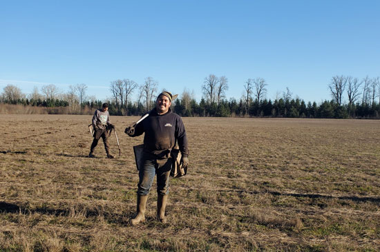 A tree planter stands smiling with a shovel over his shoulder in an open field